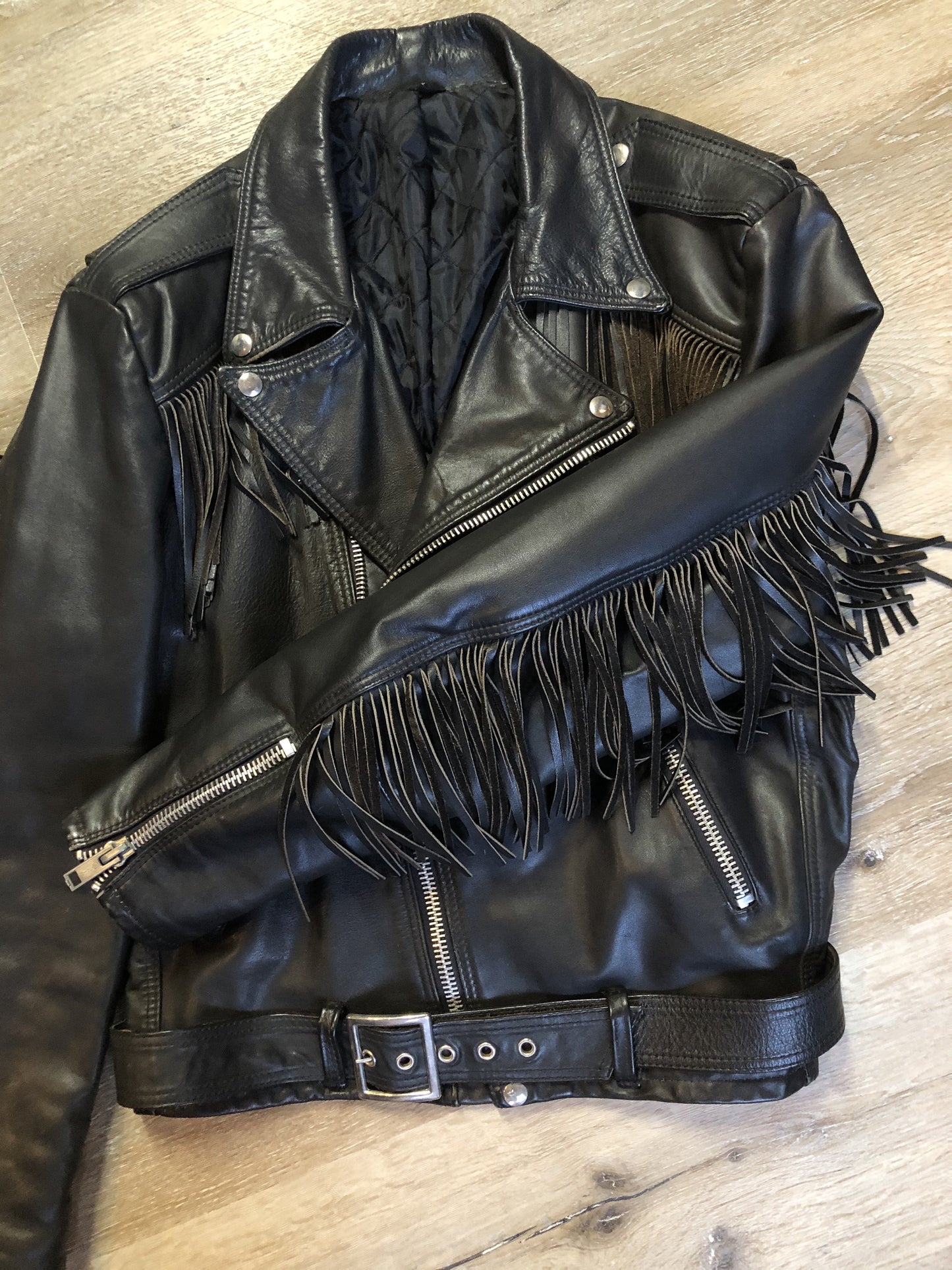 Kingspier Vintage - Action black leather motorcycle jacket with fringe detail and belt at waist. Zipper closure and zip slash pockets, quilted lining with inside pocket. Made in Canada. Size 40.