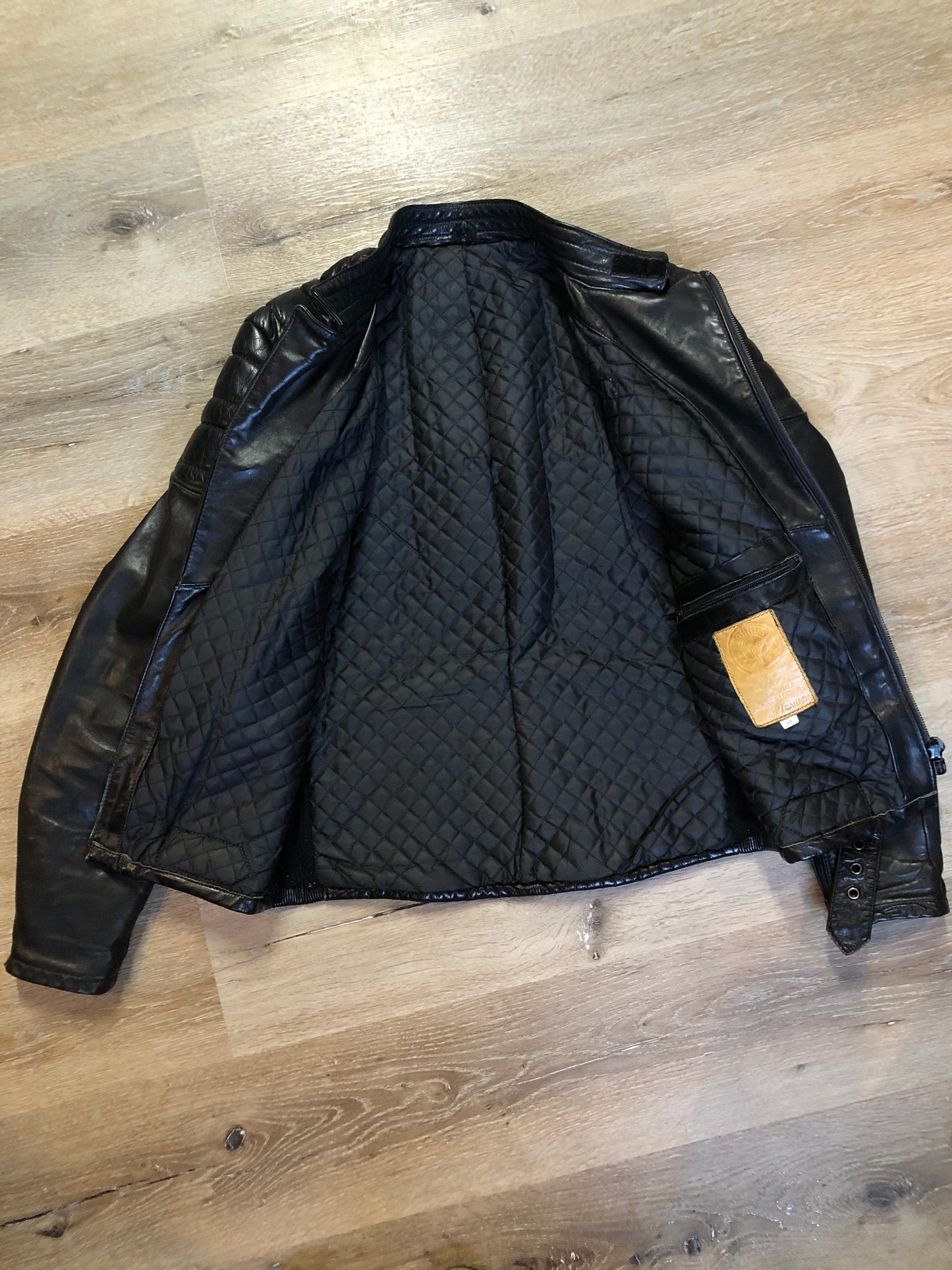 Kingspier Vintage - Taurus by Drospo black leather moto jacket with stretch detailing on the sides and a belt at the waist, zipper down the front, standing collar with Velcro strap, slash pockets and a quilted lining with inside pocket. Size 38. Made in Canada.