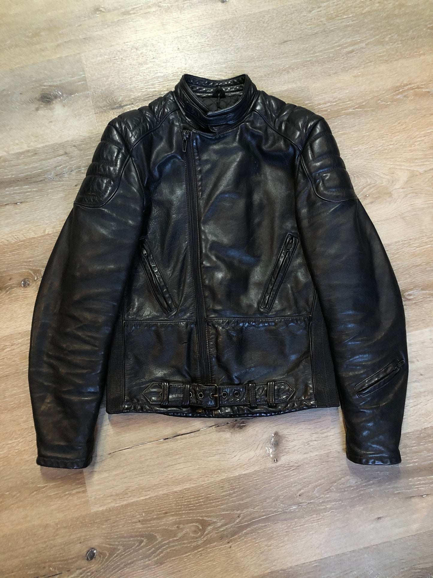 Kingspier Vintage - Taurus by Drospo black leather moto jacket with stretch detailing on the sides and a belt at the waist, zipper down the front, standing collar with Velcro strap, slash pockets and a quilted lining with inside pocket. Size 38. Made in Canada.