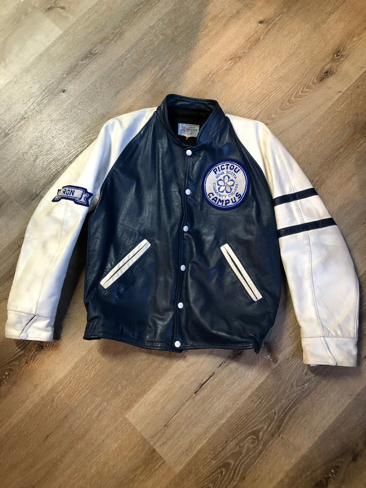 Kingspier Vintage - Vintage NSCC Pictou Campus leather varsity jacket in blue and white with snap closures, slash pockets, “Pictou Campus” embroidered on the chest, “NSCC” Embroidered on the back and “Ron” monogram embroidered on the sleeve,