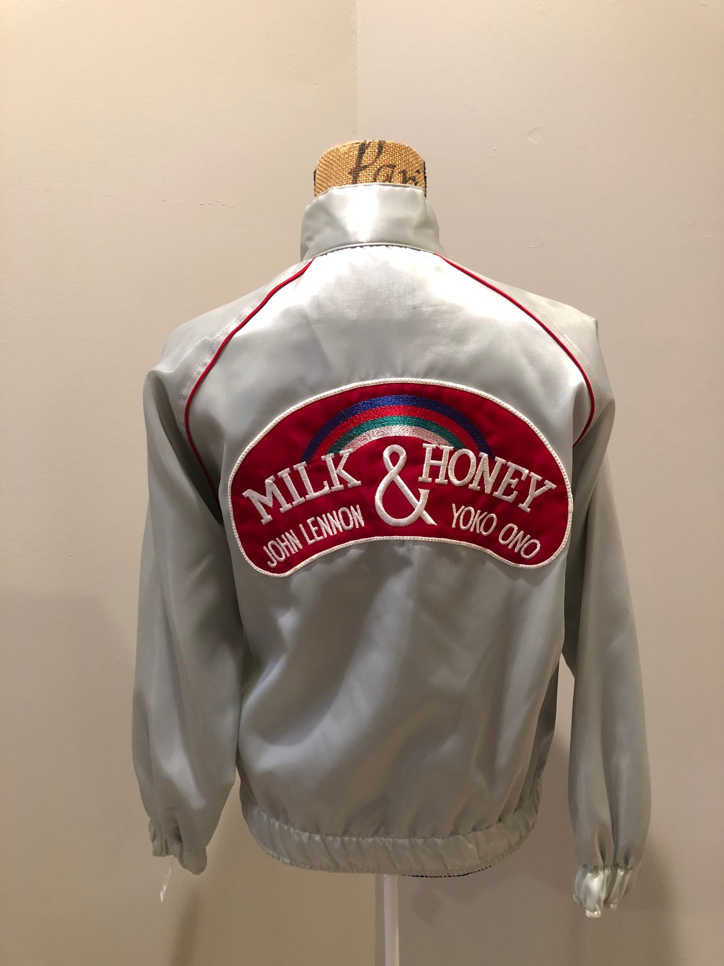 Kingspier Vintage - Milk and Honey - John Lennon and Yoko Ono Roadie Tour Jacket from the mid 1980’s. A very rare collectors piece from John Lennon’s last studio album. Jacket is light grey nylon with satin effect with two slash pockets, elastic waist and cuffs, chunky red zip up front with popper at the collar. The jacket features a star logo on the front and Milk and honey logo on the back with a rainbow. Made in the USA. Size XS.