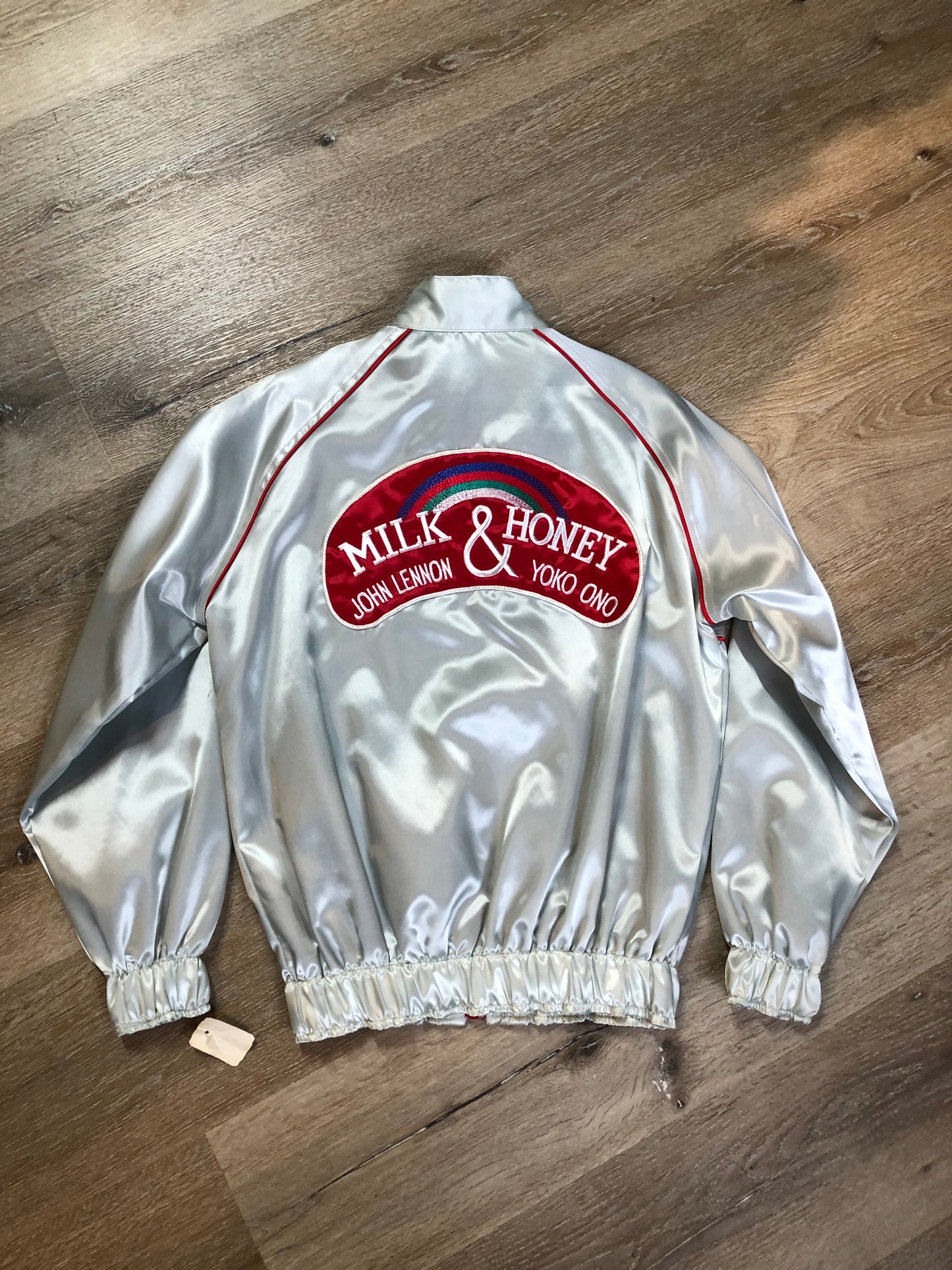 Kingspier Vintage - Milk and Honey - John Lennon and Yoko Ono Roadie Tour Jacket from the mid 1980’s. A very rare collectors piece from John Lennon’s last studio album. Jacket is light grey nylon with satin effect with two slash pockets, elastic waist and cuffs, chunky red zip up front with popper at the collar. The jacket features a star logo on the front and Milk and honey logo on the back with a rainbow. Made in the USA. Size XS.