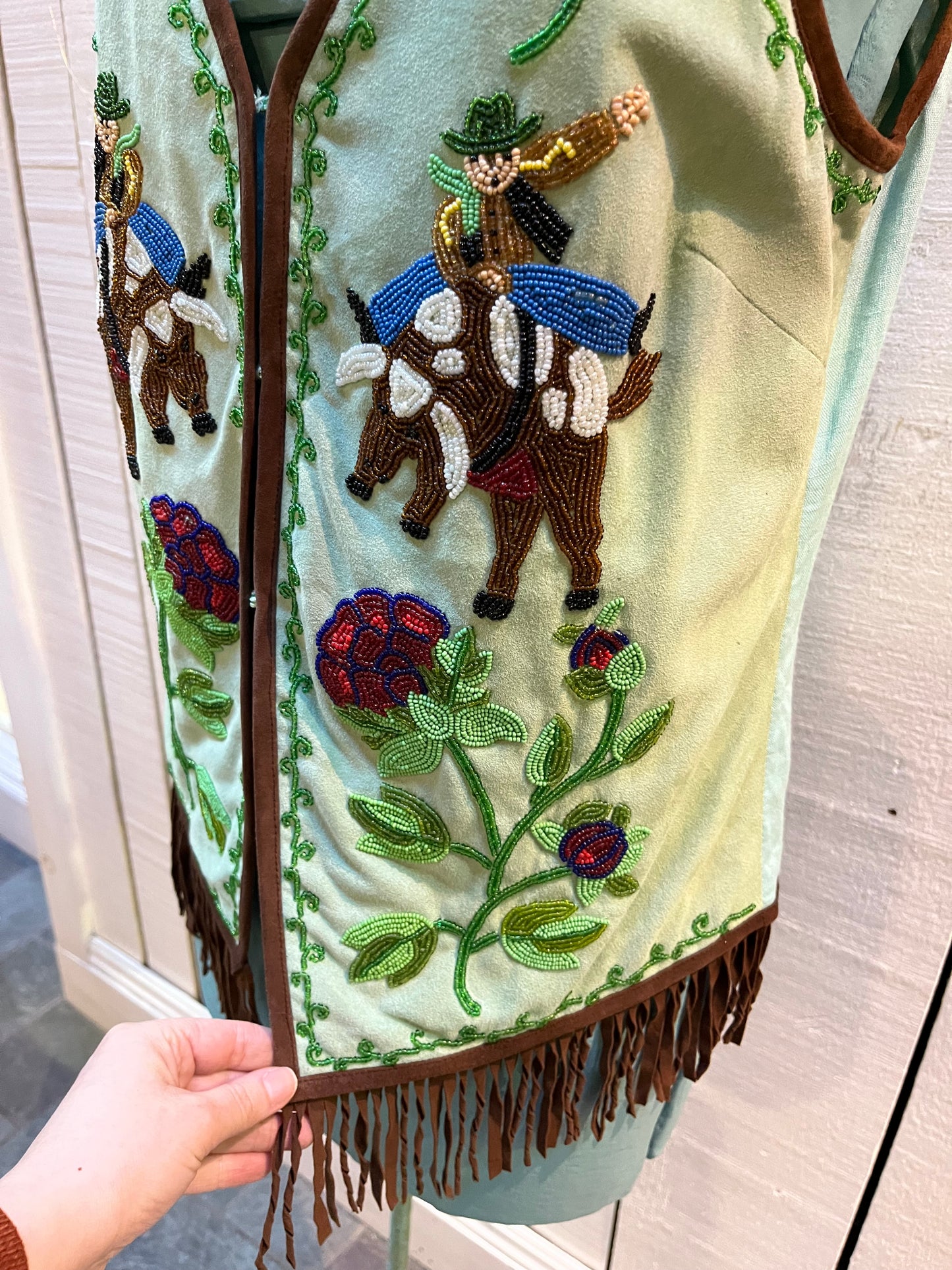 Double D Ranch light green goat suede vest with brown fringe detail, hook and eye closure, cotton/ linen back panel, satin lining and a western cowboy and desert rose design that is hand beaded.
Made in India
Size Medium