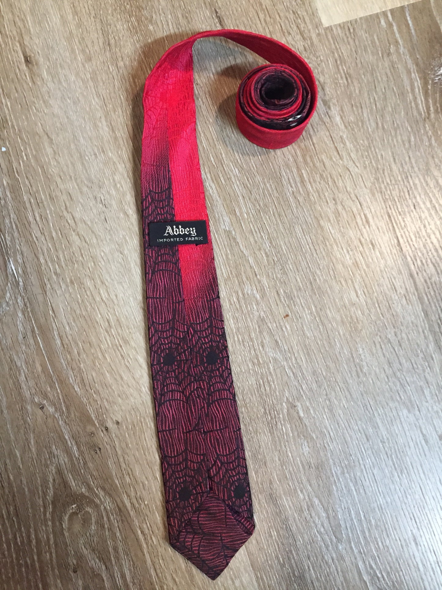 Kingspier Vintage - Abbey red tie with black spider web motif. Fibres unknown.
Length: 55”
Width: 2.5” 
This tie is in excellent condition.