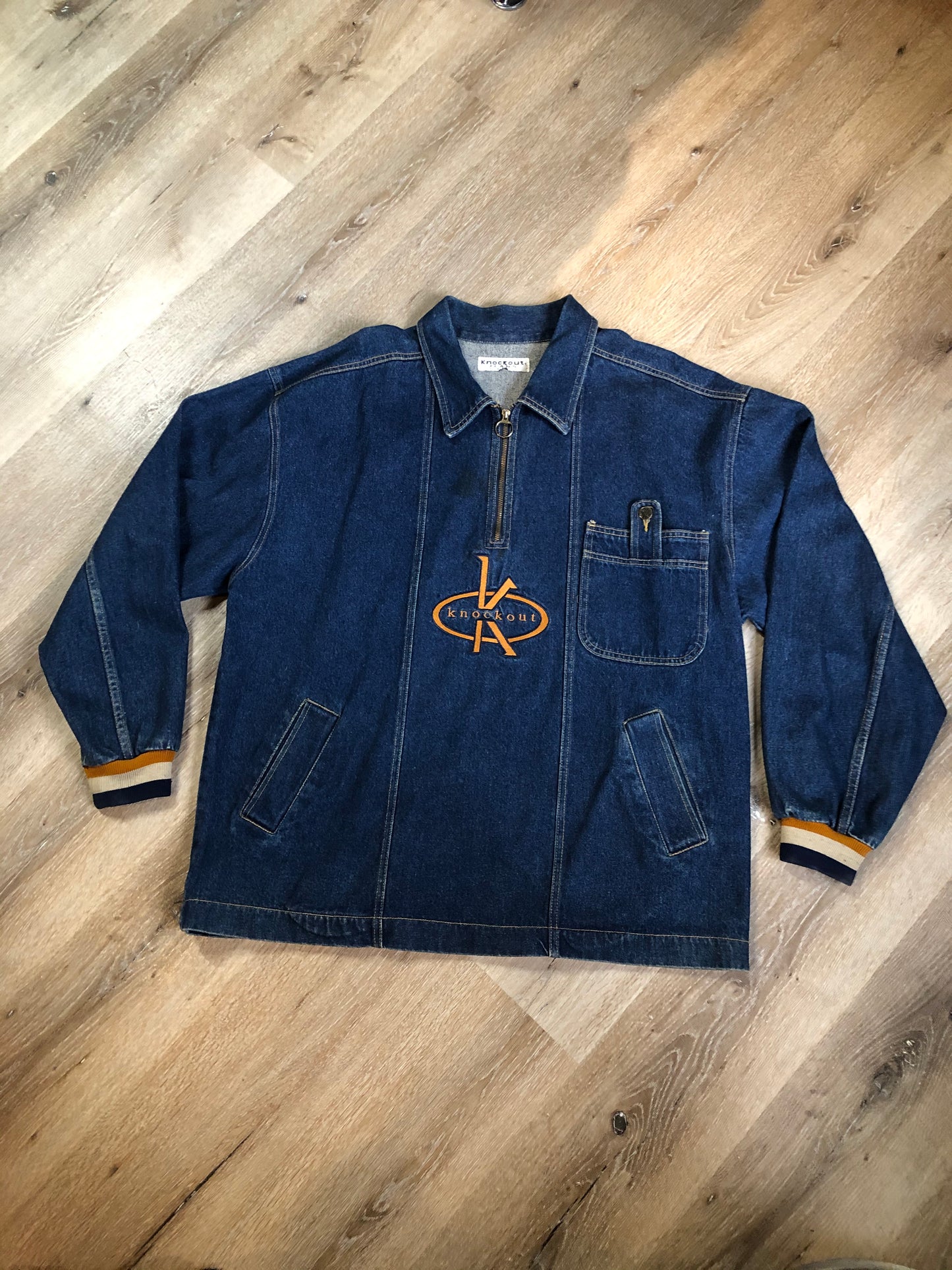 Kingspier Vintage - Vintage Knockout Jeans pullover denim jacket in a medium wash with quarter zip closure, embroidered logo on the front, knit cuffs, slash pockets, one patch pocket on the chest and a leather patch on the back. Size Xl. Made in USA