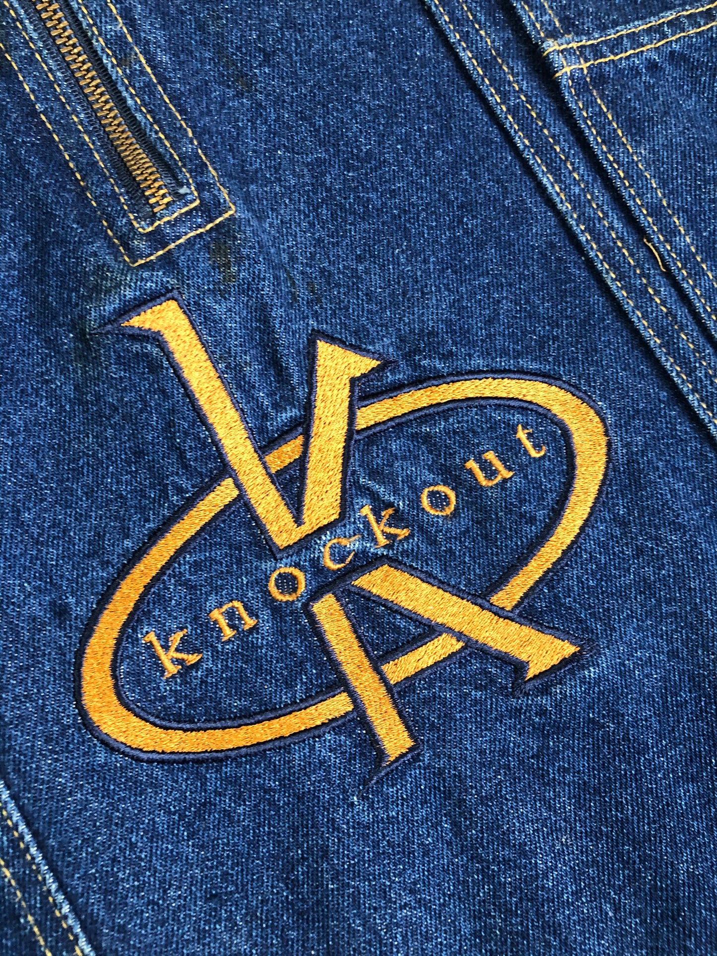 Kingspier Vintage - Vintage Knockout Jeans pullover denim jacket in a medium wash with quarter zip closure, embroidered logo on the front, knit cuffs, slash pockets, one patch pocket on the chest and a leather patch on the back. Size Xl. Made in USA