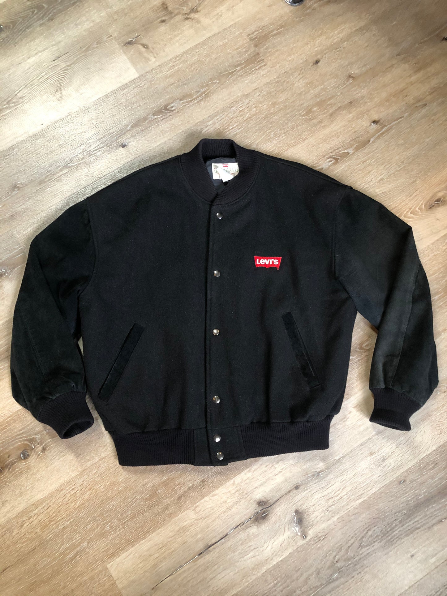 Kingspier Vintage - Vintage Levi’s black wool blend varsity jacket with Levi’s logo embroidered on the chest, “Levi’s Blue Jeans, Factory Made” with a large illustration of a factory across the back, micro suede sleeves, snap closures, slash pockets, polyester lining, one inside pocket and knit trim. Made in Canada. Size medium.