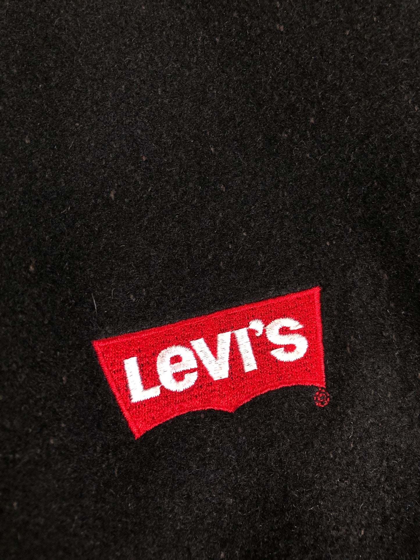 Kingspier Vintage - Vintage Levi’s black wool blend varsity jacket with Levi’s logo embroidered on the chest, “Levi’s Blue Jeans, Factory Made” with a large illustration of a factory across the back, micro suede sleeves, snap closures, slash pockets, polyester lining, one inside pocket and knit trim. Made in Canada. Size medium.