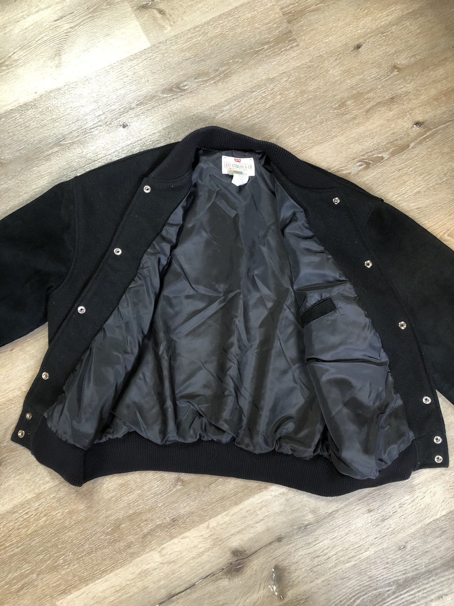 Kingspier Vintage - Vintage Levi’s black wool blend varsity jacket with Levi’s logo embroidered on the chest, “Levi’s Blue Jeans, Factory Made” with a large illustration of a factory across the back, micro suede sleeves, snap closures, slash pockets, polyester lining, one inside pocket and knit trim. Made in Canada. Size medium.