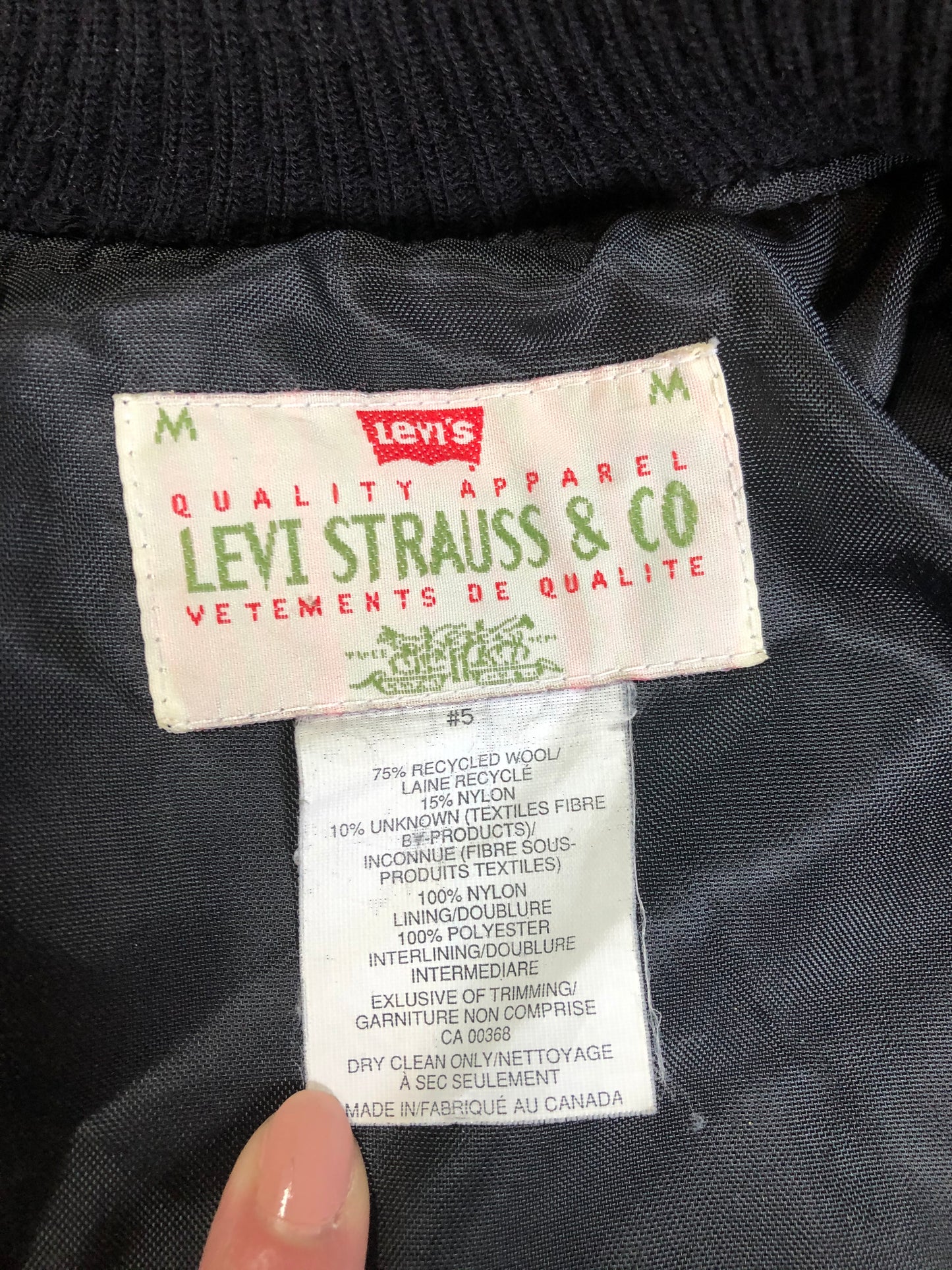 Kingspier Vintage - Vintage Levi’s black wool blend varsity jacket with Levi’s logo embroidered on the chest, “Levi’s Blue Jeans, Factory Made” with a large illustration of a factory across the back, micro suede sleeves, snap closures, slash pockets, polyester lining, one inside pocket and knit trim. Made in Canada. Size medium.