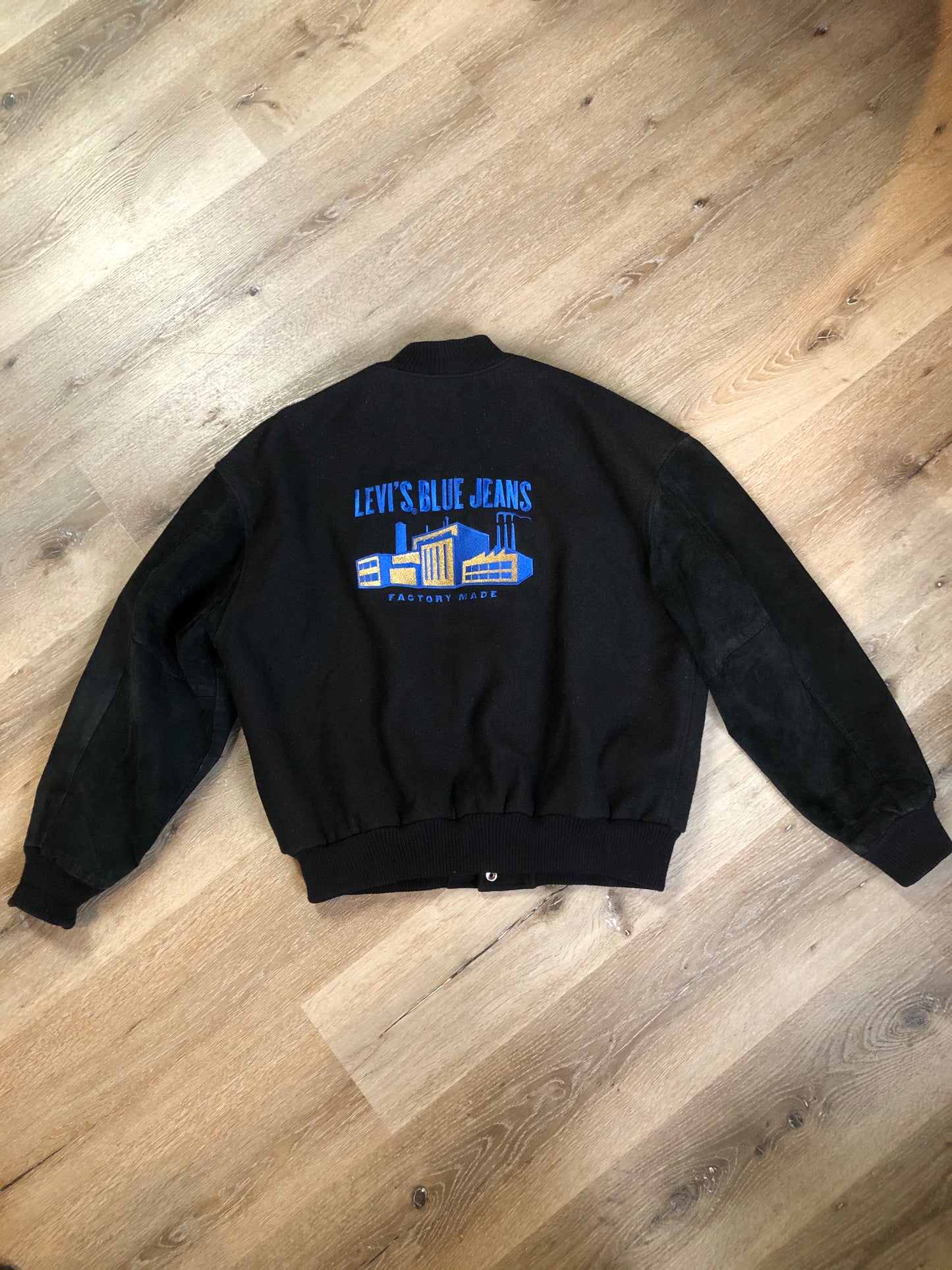 Kingspier Vintage - Vintage Levi’s black wool blend varsity jacket with Levi’s logo embroidered on the chest, “Levi’s Blue Jeans, Factory Made” with a large illustration of a factory across the back, micro suede sleeves, snap closures, slash pockets, polyester lining, one inside pocket and knit trim. Made in Canada. Size medium.