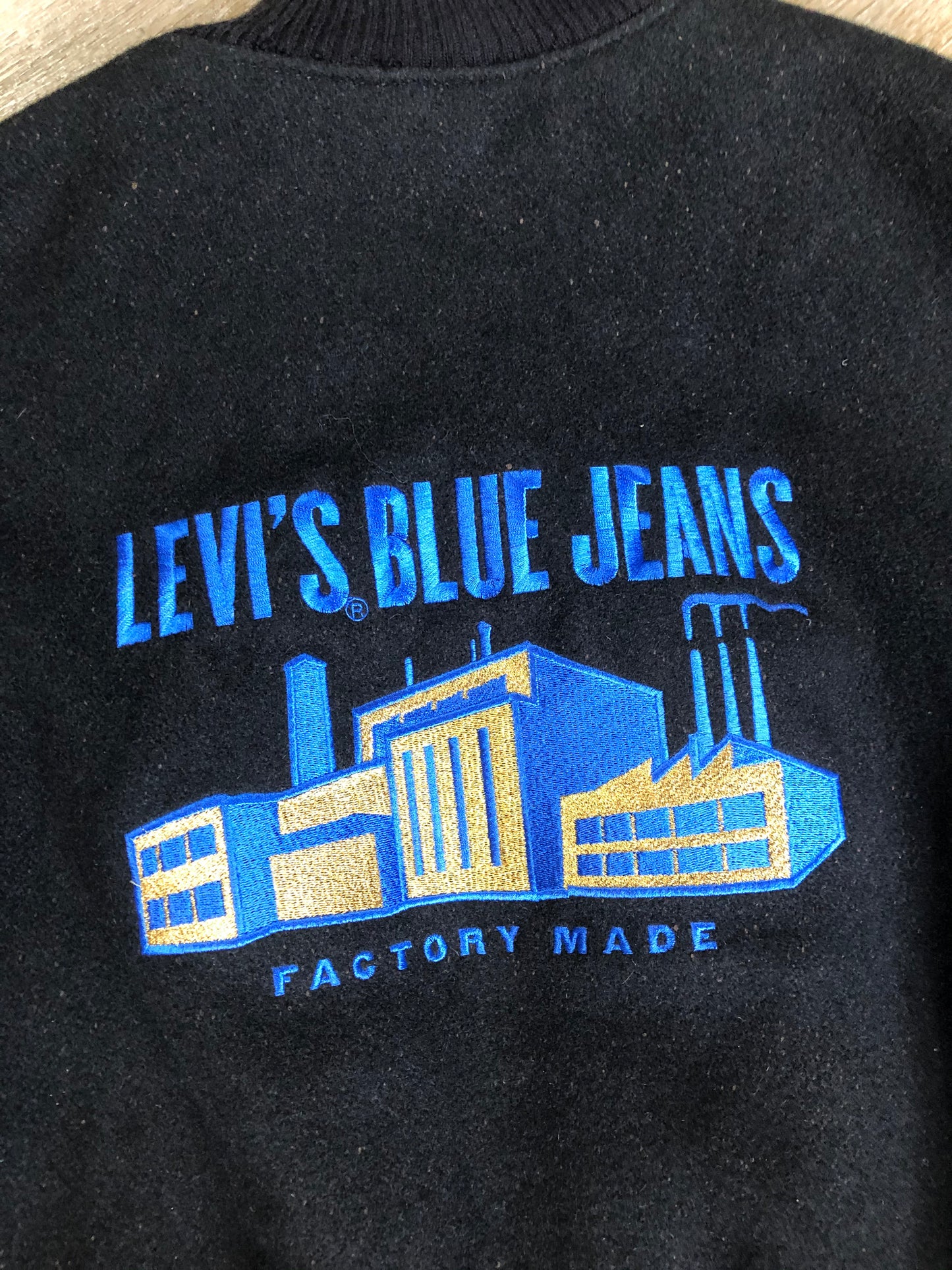 Kingspier Vintage - Vintage Levi’s black wool blend varsity jacket with Levi’s logo embroidered on the chest, “Levi’s Blue Jeans, Factory Made” with a large illustration of a factory across the back, micro suede sleeves, snap closures, slash pockets, polyester lining, one inside pocket and knit trim. Made in Canada. Size medium.