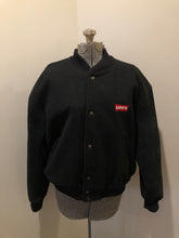 Load image into Gallery viewer, Kingspier Vintage - Vintage Levi’s black wool blend varsity jacket with Levi’s logo embroidered on the chest, “Levi’s Blue Jeans, Factory Made” with a large illustration of a factory across the back, micro suede sleeves, snap closures, slash pockets, polyester lining, one inside pocket and knit trim. Made in Canada. Size medium.
