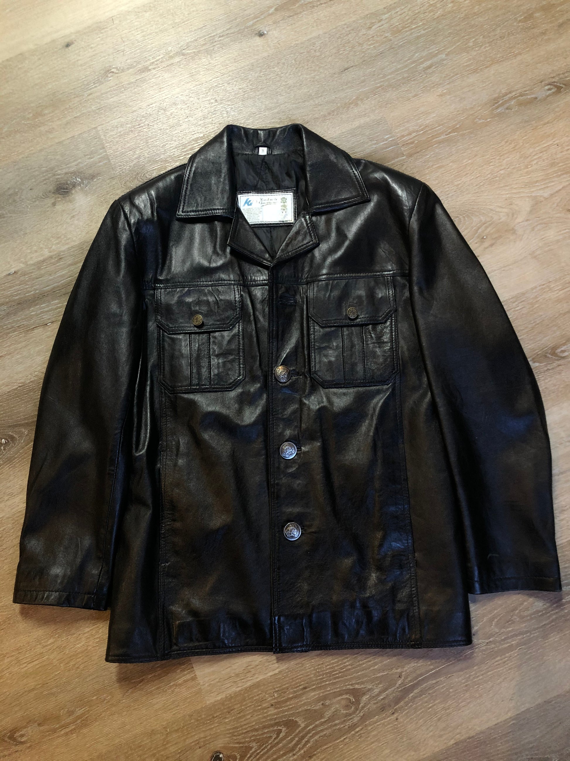 Kingspier Vintage - Kashmir Garments black leather jacket features two flap pockets on the chest with hidden chain detail underneath and skull button closures. Size medium.