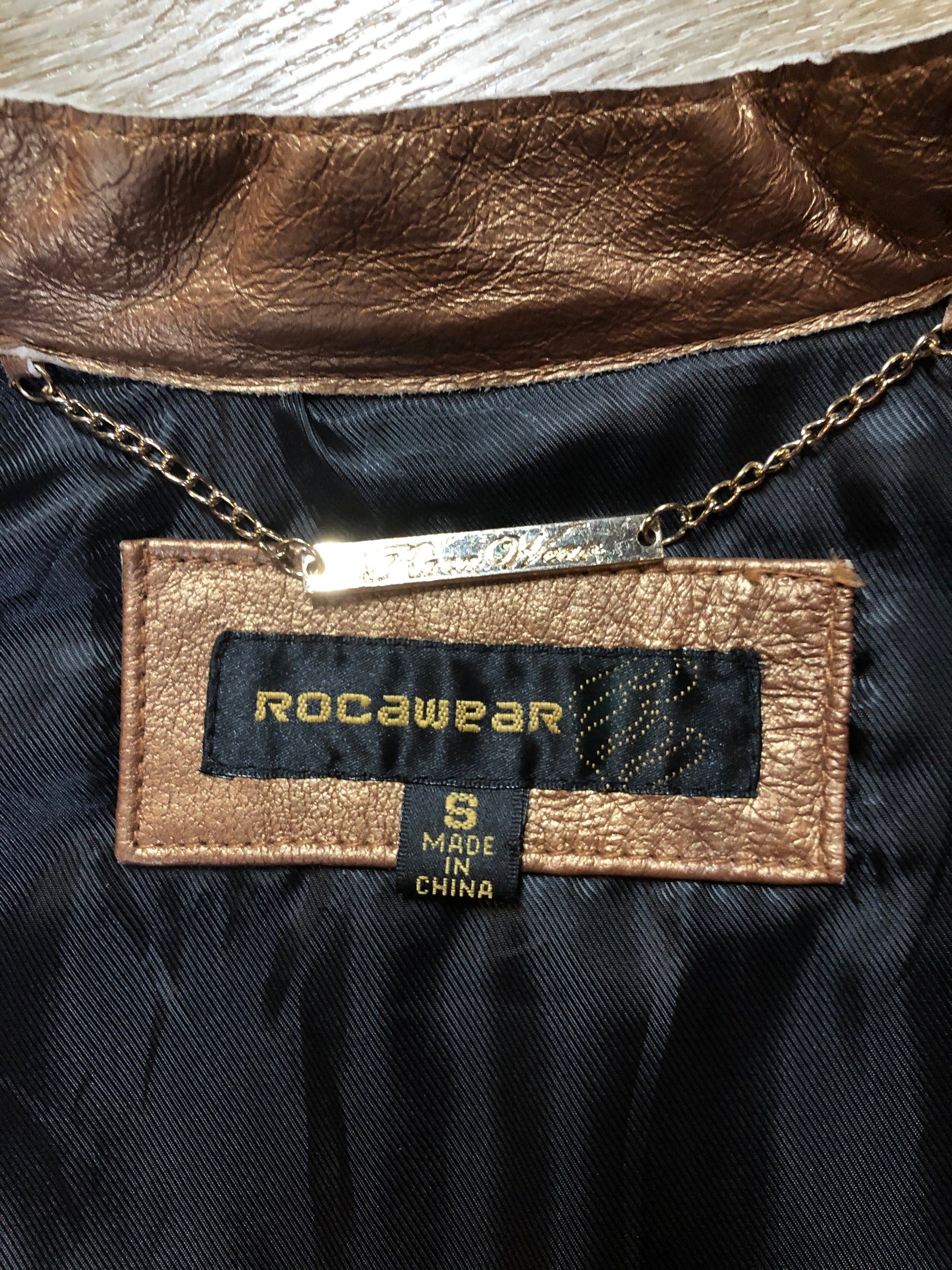 Kingspier Vintage - Rocawear metallic gold leather moto jacket with “RW” detail zippers, chain lace-up detail on sides and “ROCAWEAR” written in chain across the back. There is a front zipper, two horizontal zip pockets on the chest, two snap closures on the stand up collar and a pocket on the inside. Size small.