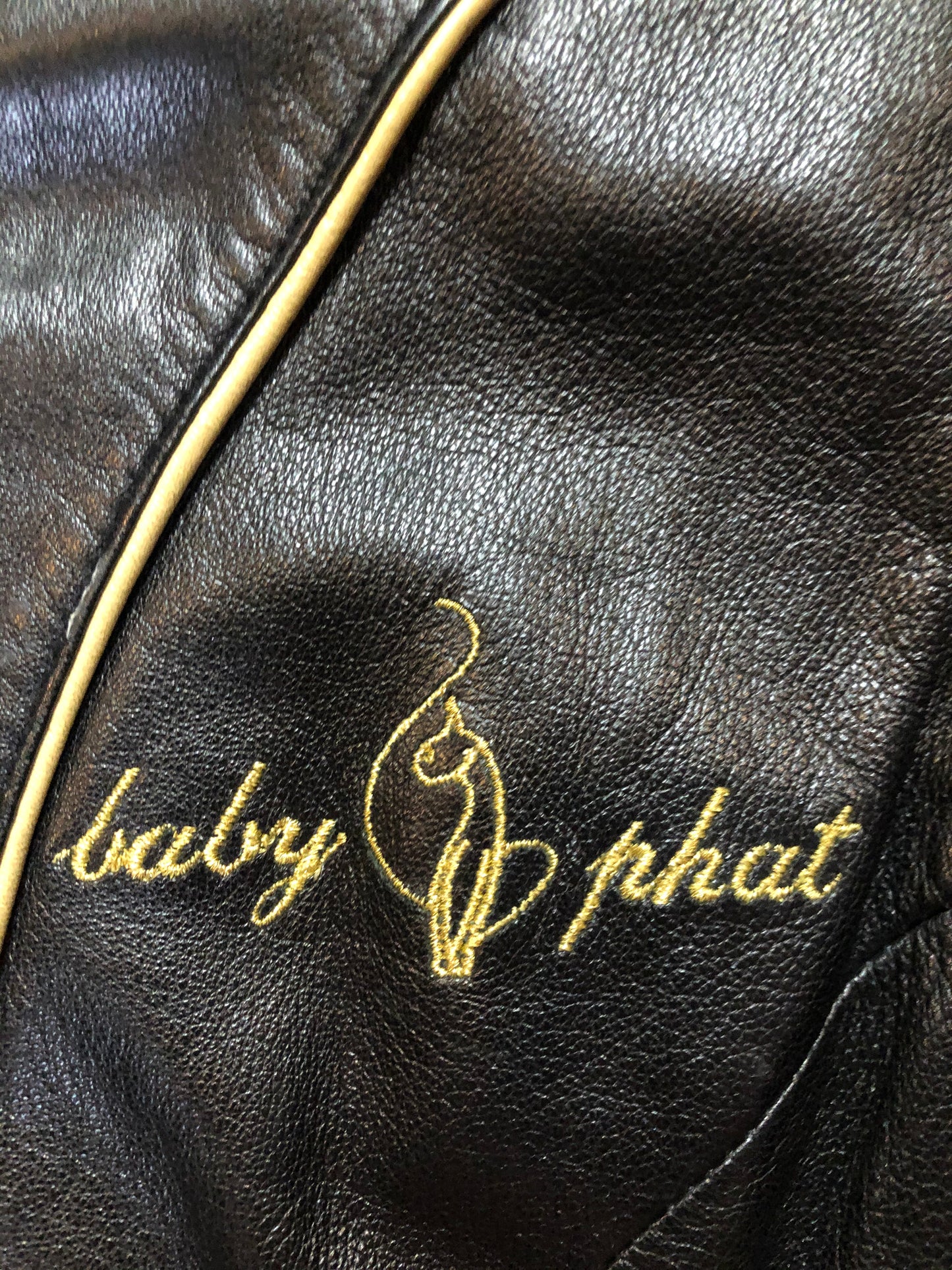 Kingspier Vintage - Baby Phat black leather moto jacket with gold piping and “Baby Phat” embroidered on the chest, front zipper and two vertical zip pockets. A pattern is stitched into the elbows and “Baby Phat” is stitched across the back. Size small.