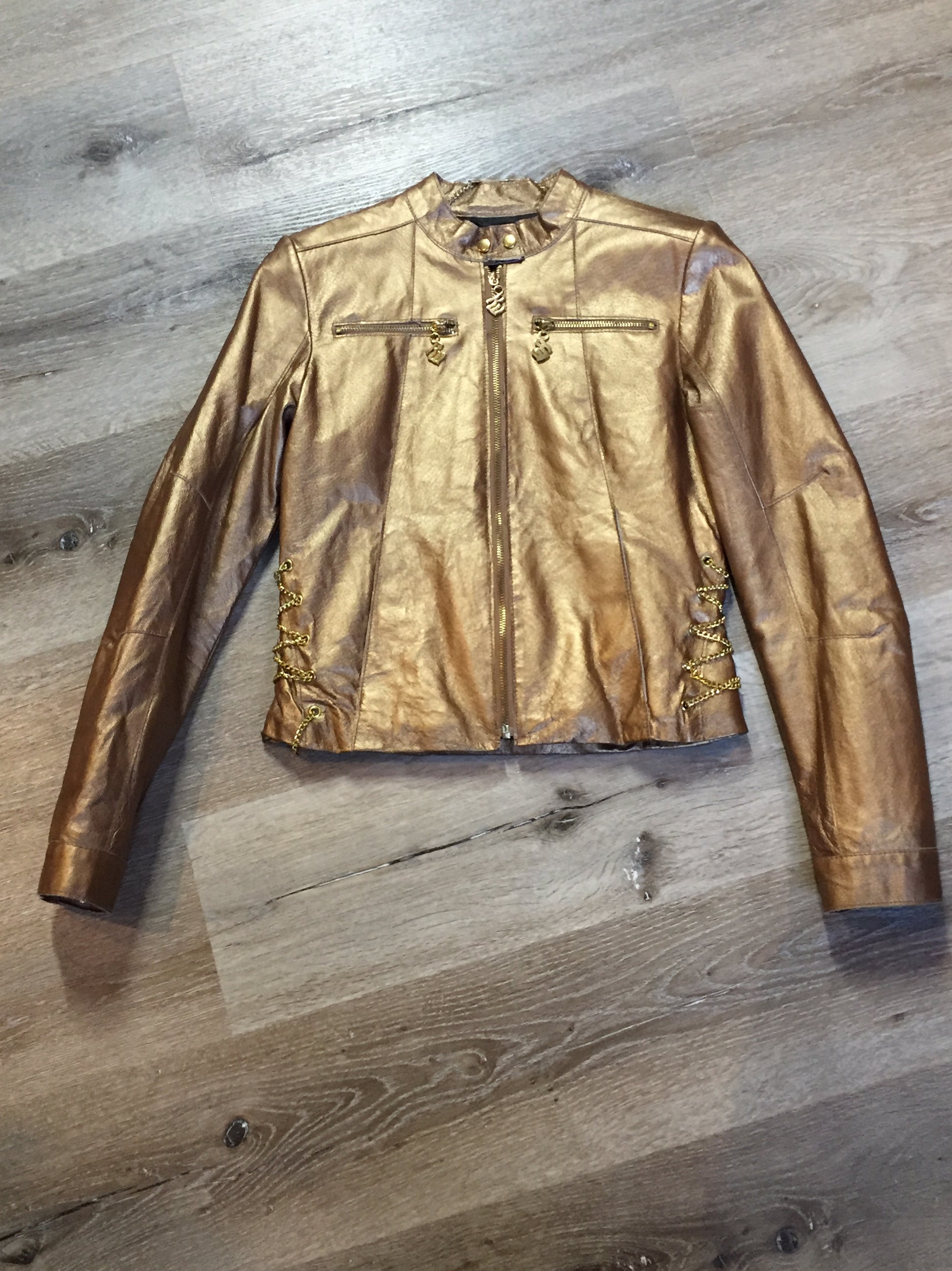 Kingspier Vintage - Rocawear metallic gold leather moto jacket with “RW” detail zippers, chain lace-up detail on sides and “ROCAWEAR” written in chain across the back. There is a front zipper, two horizontal zip pockets on the chest, two snap closures on the stand up collar and a pocket on the inside. Size small.