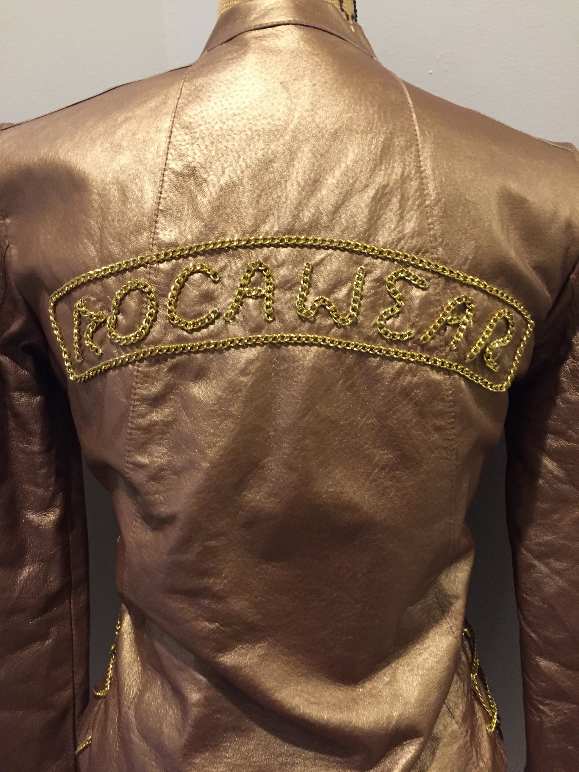 Kingspier Vintage - Rocawear metallic gold leather moto jacket with “RW” detail zippers, chain lace-up detail on sides and “ROCAWEAR” written in chain across the back. There is a front zipper, two horizontal zip pockets on the chest, two snap closures on the stand up collar and a pocket on the inside. Size small.