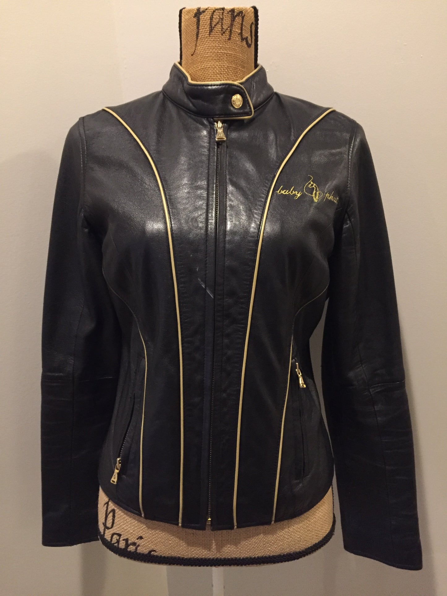 Kingspier Vintage - Baby Phat black leather moto jacket with gold piping and “Baby Phat” embroidered on the chest, front zipper and two vertical zip pockets. A pattern is stitched into the elbows and “Baby Phat” is stitched across the back. Size small.