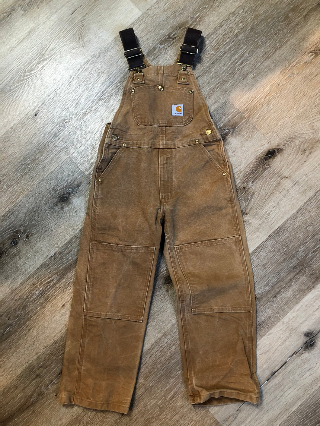 Kingspier Vintage - Kids Carhartt Sleeveless cotton canvas overalls in tan brown with Carhartt logo. Overalls feature adjustable shoulder straps with buckle fastening, patch pocket in the chest and pockets in the front and back, overalls button at the waist and have reinforced knees. Size childs medium.