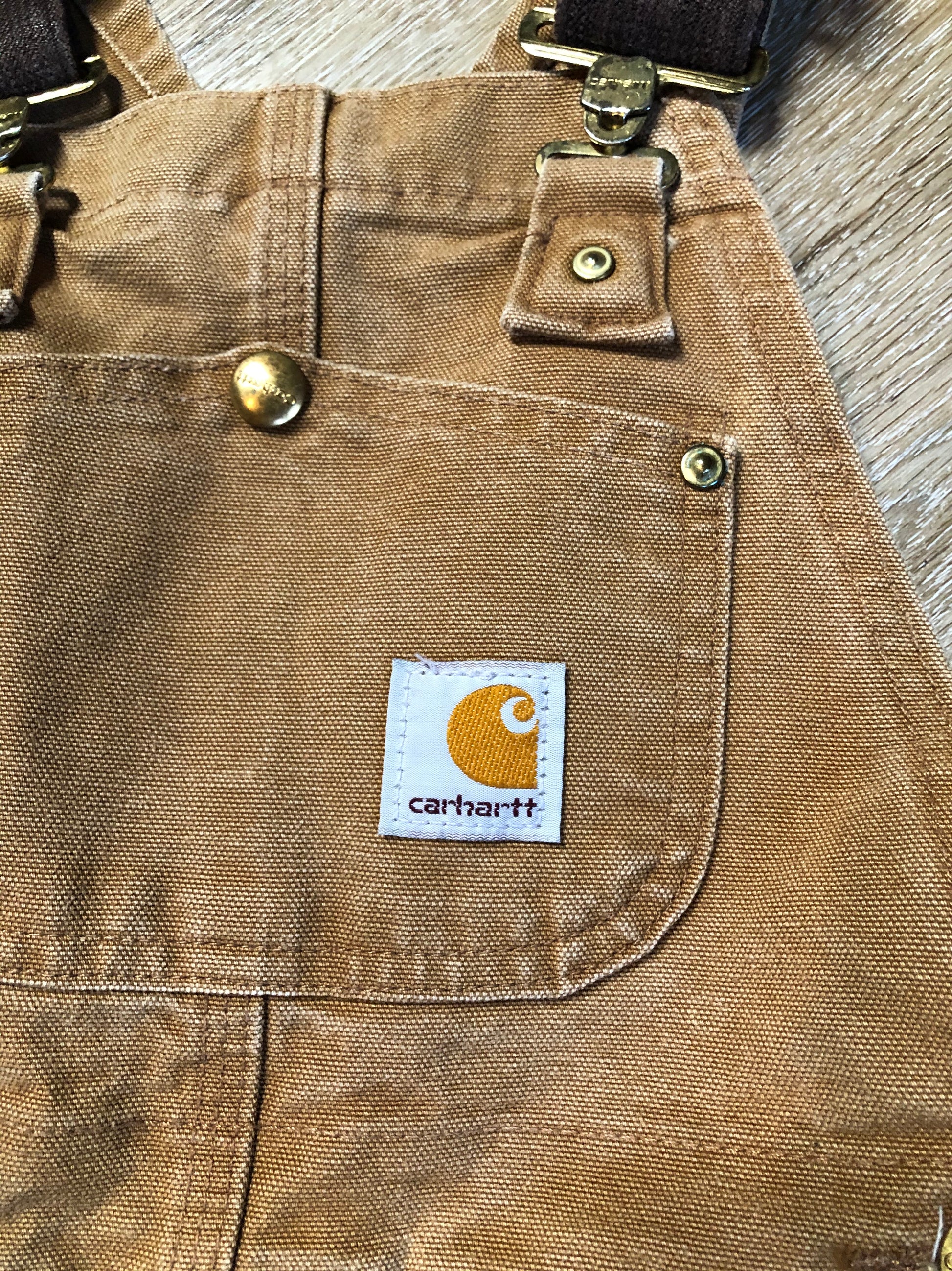 Kingspier Vintage - Kids Carhartt Sleeveless cotton canvas overalls in tan brown with Carhartt logo. Overalls feature adjustable shoulder straps with buckle fastening, patch pocket in the chest and pockets in the front and back, overalls button at the waist and have reinforced knees. Size childs medium.