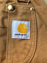 Load image into Gallery viewer, Kingspier Vintage - Carhartt Sleeveless duck canvas overalls in tan brown with Carhartt logo. Overalls feature adjustable shoulder straps with buckle fastening, hammer loop, patch pocket on the chest, side utility pockets, pockets in the front and back, overalls button at the waist and have reinforced knees. Size 32X30.
