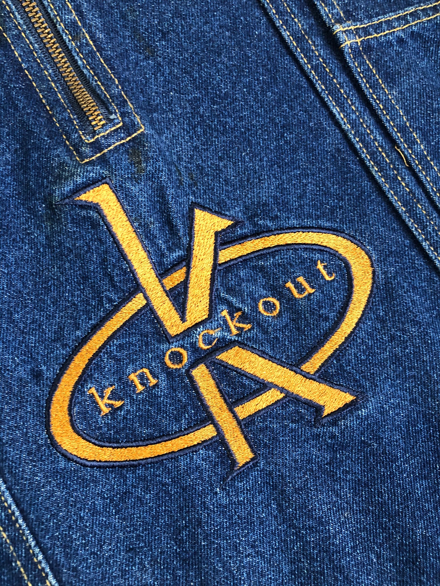 Kingspier Vintage - Vintage Knockout Jeans pullover denim jacket in a medium wash with quarter zip closure, embroidered logo on the front, knit cuffs, slash pockets, one patch pocket on the chest and a leather patch on the back. Size Xl. Made in USA
