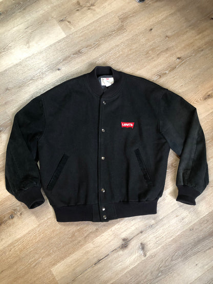 Kingspier Vintage - Vintage Levi’s black wool blend varsity jacket with Levi’s logo embroidered on the chest, “Levi’s Blue Jeans, Factory Made” with a large illustration of a factory across the back, micro suede sleeves, snap closures, slash pockets, polyester lining, one inside pocket and knit trim. Made in Canada. Size medium.
