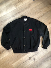 Load image into Gallery viewer, Kingspier Vintage - Vintage Levi’s black wool blend varsity jacket with Levi’s logo embroidered on the chest, “Levi’s Blue Jeans, Factory Made” with a large illustration of a factory across the back, micro suede sleeves, snap closures, slash pockets, polyester lining, one inside pocket and knit trim. Made in Canada. Size medium.

