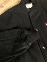 Load image into Gallery viewer, Kingspier Vintage - Vintage Levi’s black wool blend varsity jacket with Levi’s logo embroidered on the chest, “Levi’s Blue Jeans, Factory Made” with a large illustration of a factory across the back, micro suede sleeves, snap closures, slash pockets, polyester lining, one inside pocket and knit trim. Made in Canada. Size medium.

